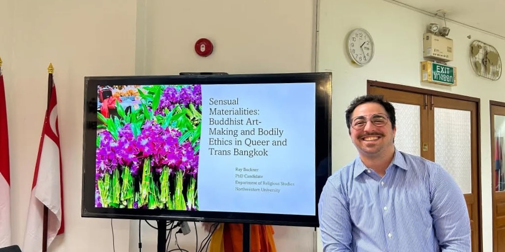 Buckner posing in front of his research presentation at a monastic university. For this presentation, he spoke with Buddhist monastics to understand their perspectives about queer and transgender art. (Bangkok, Thailand)
