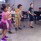 A group of children dance in a classroom guided by an instructor.