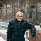 V.S. Subrahmanian is smiling and leaning one arm on a brick pillar. He is wearing a dark plaid overcoat, a black jacket , a red scarf, and black gloves.